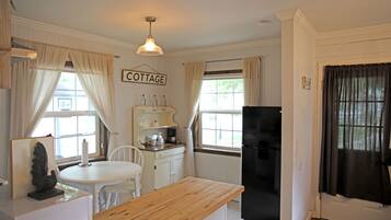 A compact kitchen with a wooden countertop, white cabinetry, and a black refrigerator. A 'COTTAGE' sign hangs on the wall, and there are two windows with curtains.