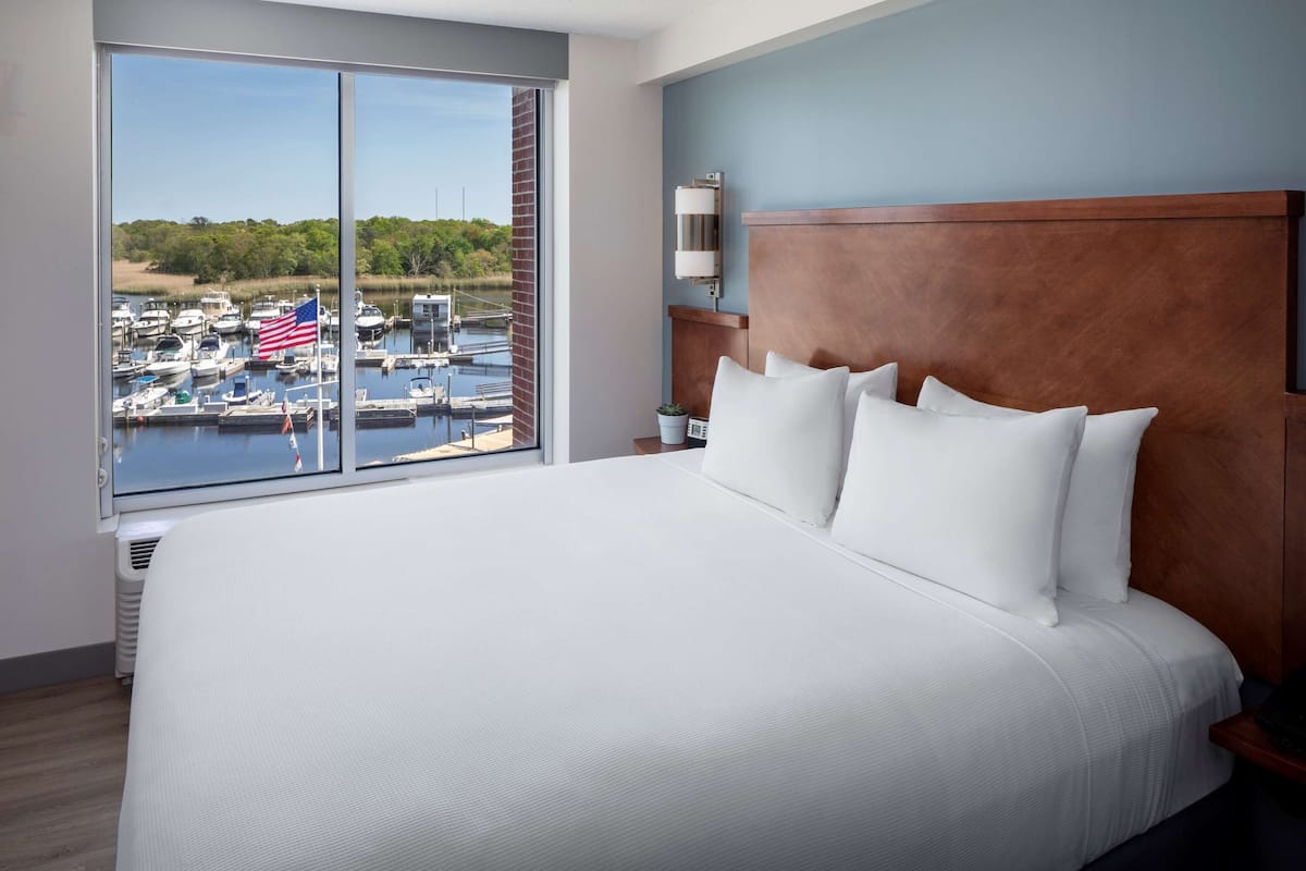 A hotel room with a large bed, a wooden headboard, and a view of a marina with boats and an American flag.