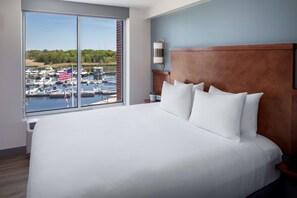 A hotel room with a large bed, a wooden headboard, and a view of a marina with boats and an American flag. - Hyatt Place Long Island East End (Riverhead)