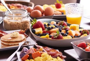 A breakfast table with pancakes, scrambled eggs, bacon, and a fruit bowl containing strawberries, blueberries, and cantaloupe. - Hyatt Place Long Island East End (Riverhead)