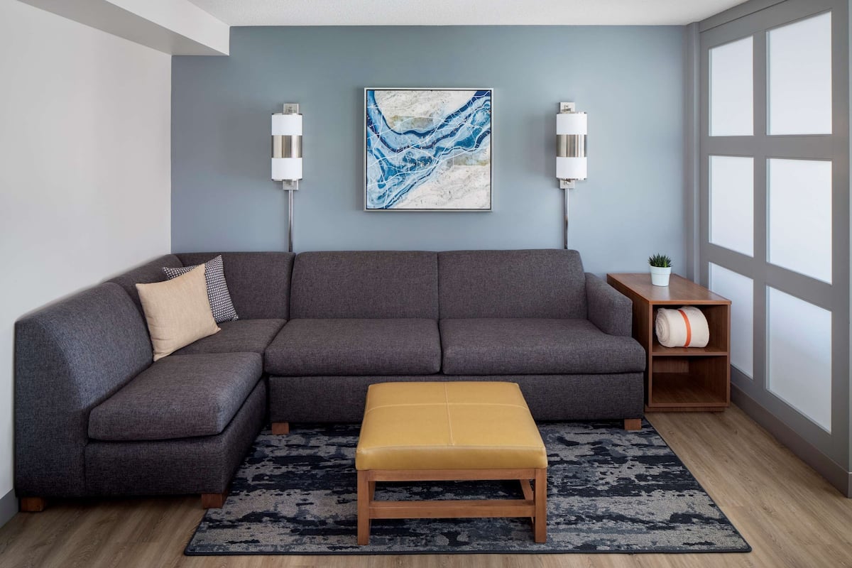A modern living room with a gray sectional sofa, a mustard-colored ottoman, a blue abstract painting, and two wall-mounted lamps.
