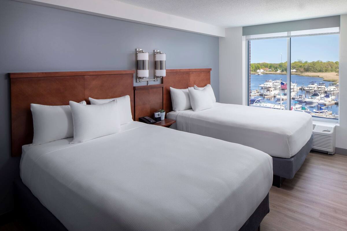 A hotel room with two beds, a wooden headboard, a nightstand, a phone, and a large window with a marina view.