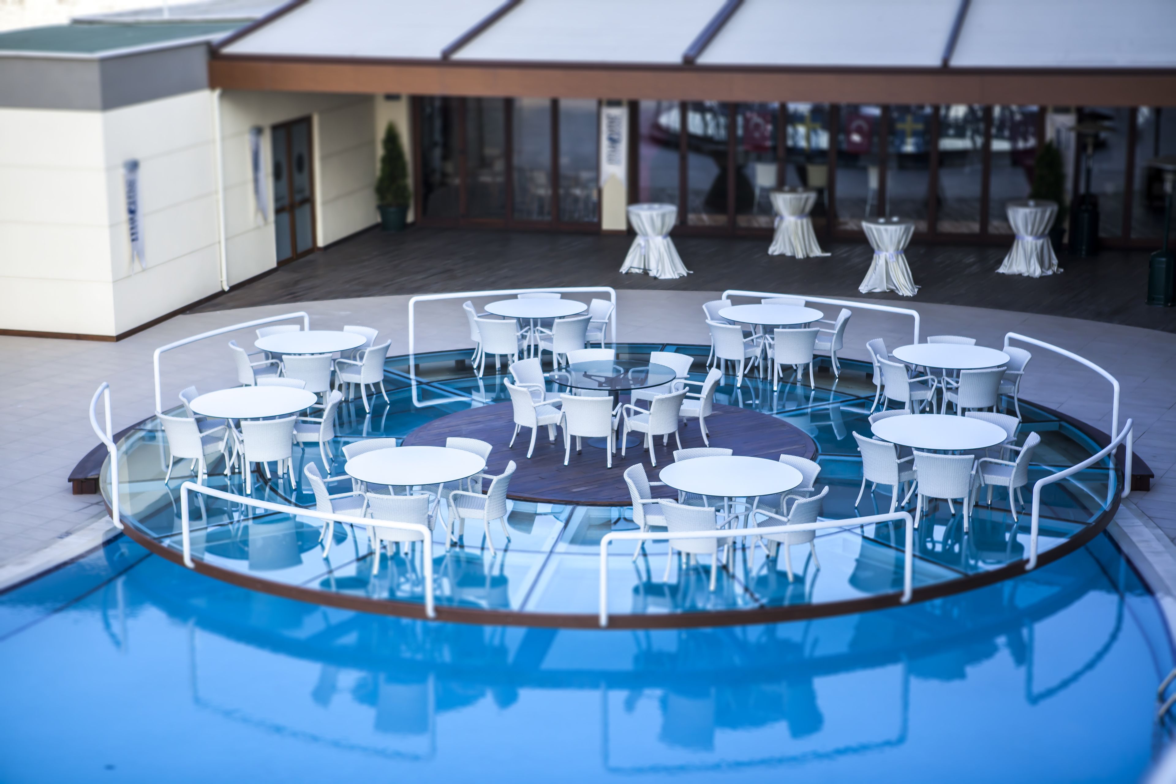 Zona de piscina con varias mesas redondas y sillas, delimitada por una barrera de cristal.