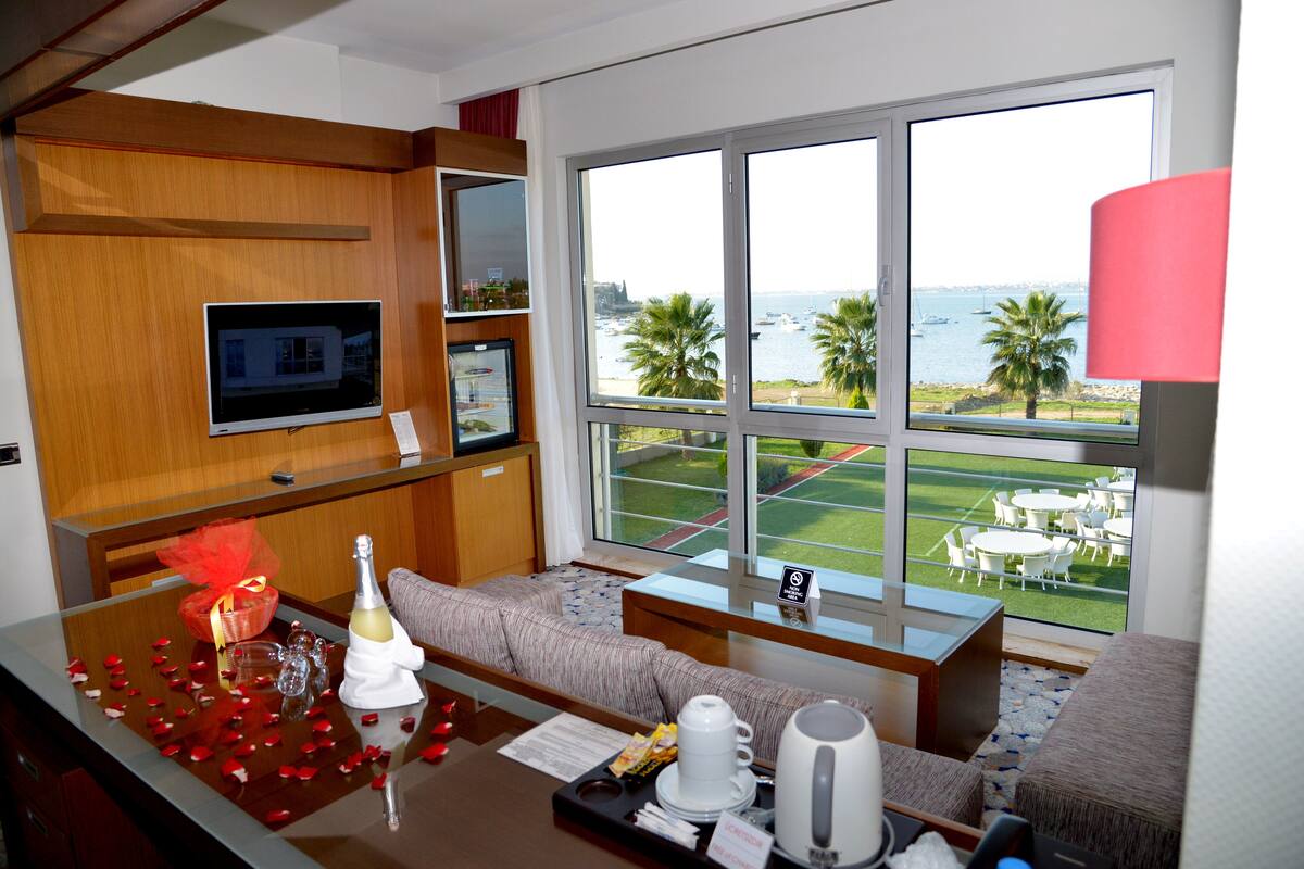 Habitación de hotel con un ventanal grande que ofrece vistas al mar y palmeras. Hay un sofá, una mesita con una botella y vasos, y un televisor montado en la pared.