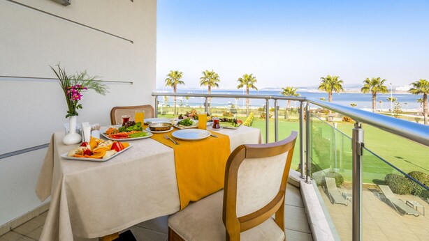 Un balcon avec une table préparée pour le petit-déjeuner, offrant une vue sur la côte, avec des palmiers et la mer.