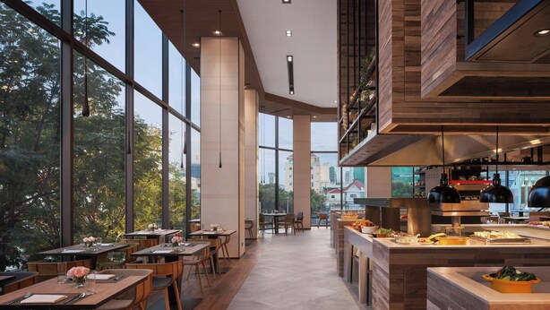 A modern restaurant with large windows, wooden tables, and a central kitchen area. - Courtyard by Marriott Phnom Penh (Phnom Penh)
