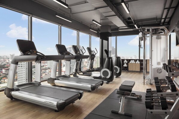 A gym with treadmills, elliptical machines, and weight benches, featuring large windows with a city view.