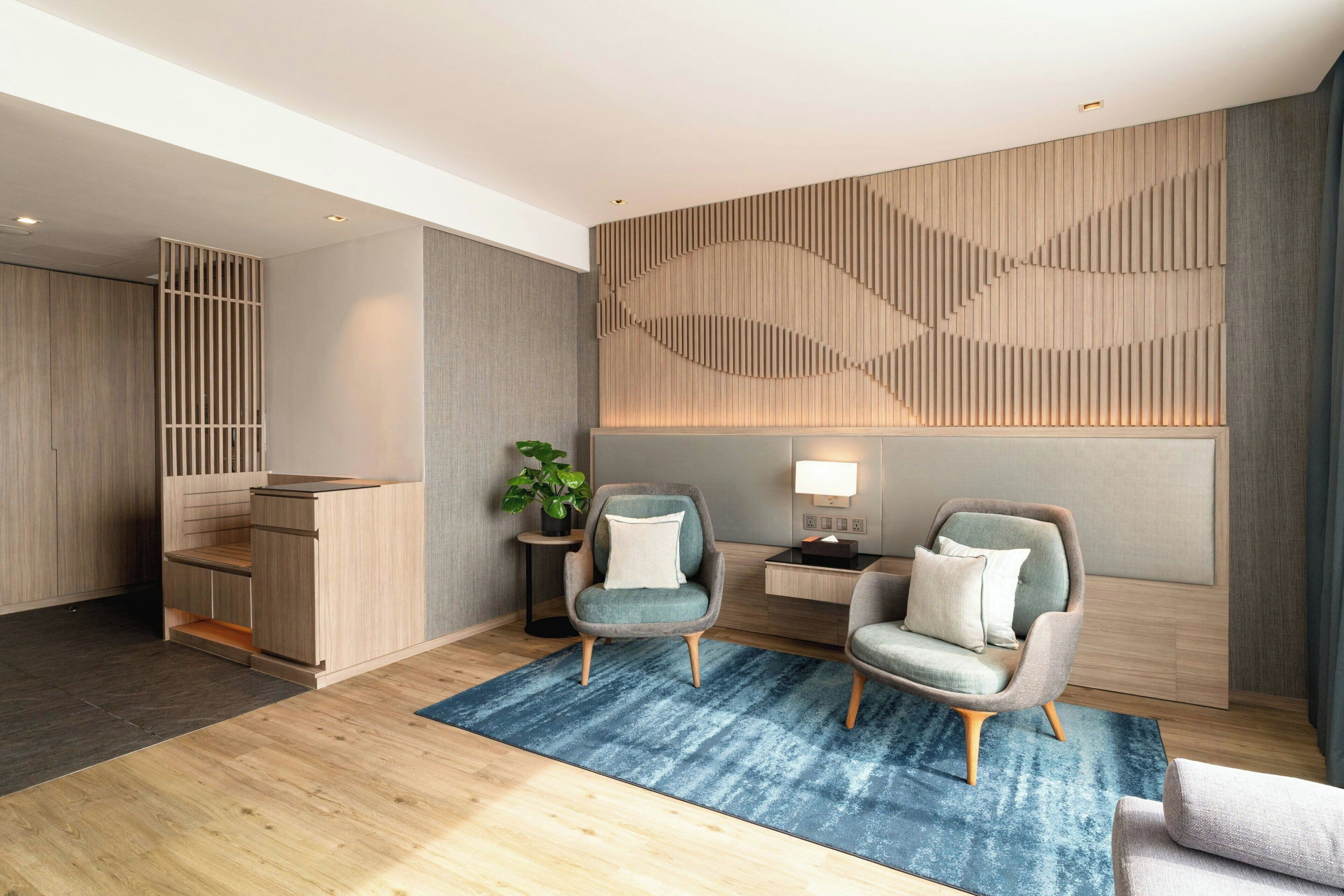 Une chambre d’hôtel moderne avec un comptoir d’accueil en bois, deux fauteuils, un tapis bleu et un luminaire fixé au mur.
