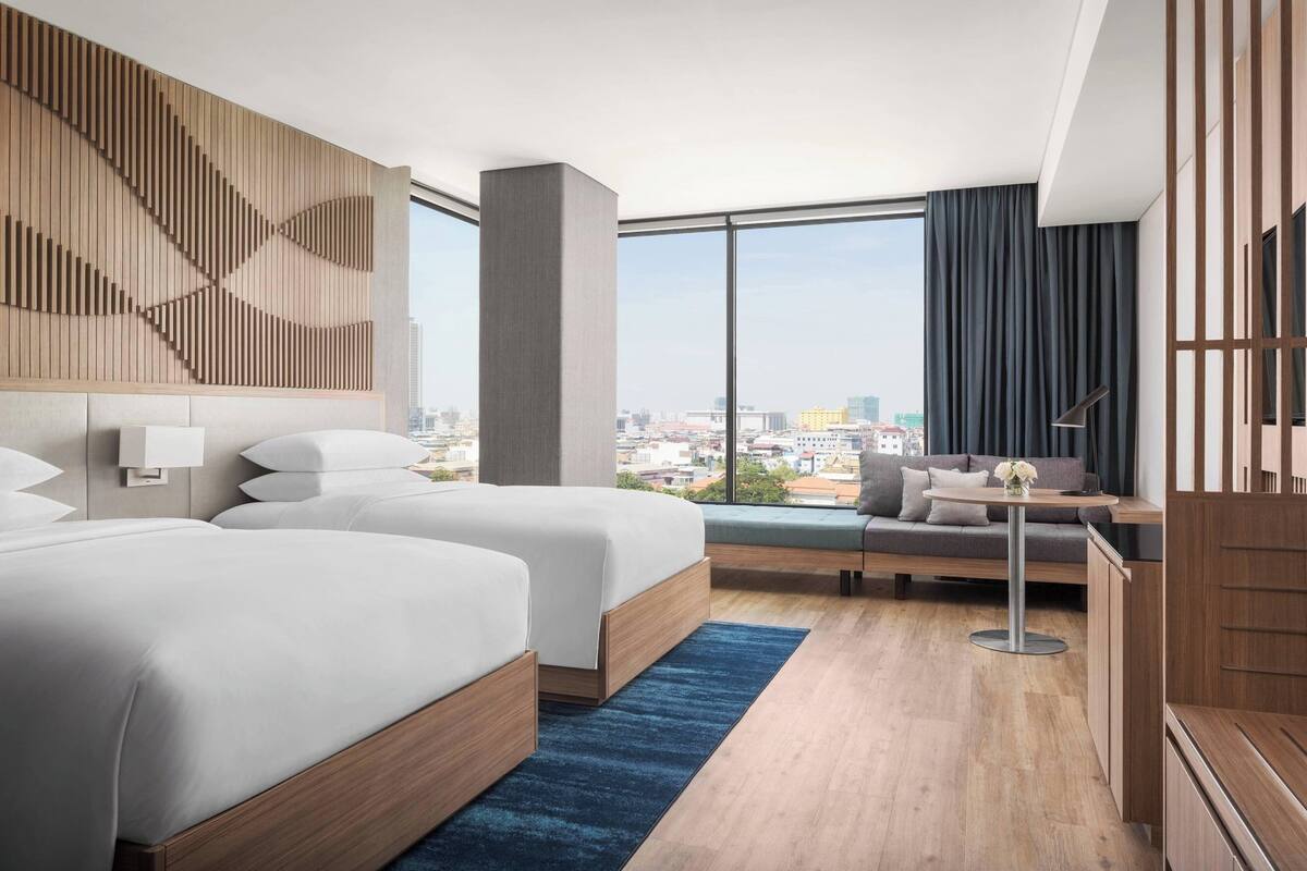 Una habitación de hotel moderna con dos camas, un ventanal con vistas a la ciudad, un panel decorativo de madera y un rincón de estar con una mesa y cojines.