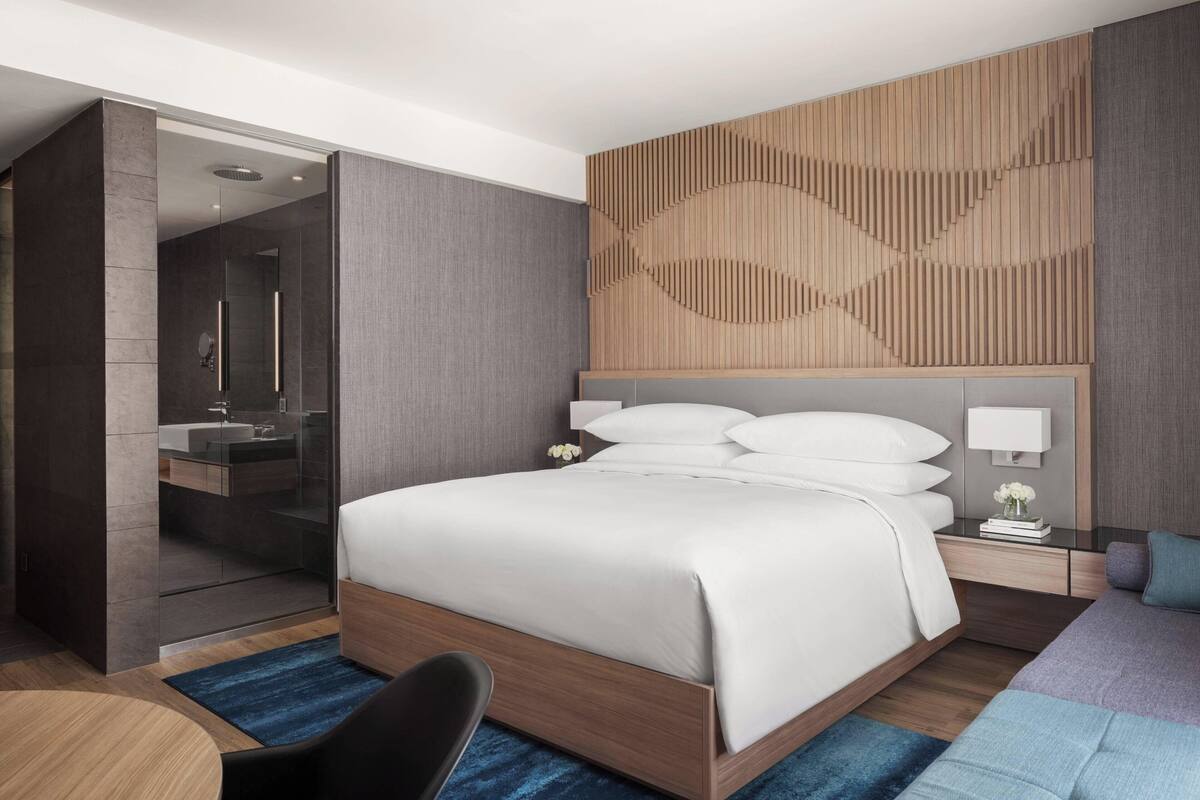 Habitación de hotel moderna con una cama grande, cabina de ducha de vidrio, tablero de cabecera de madera, mesita de noche con lámpara y alfombra azul.