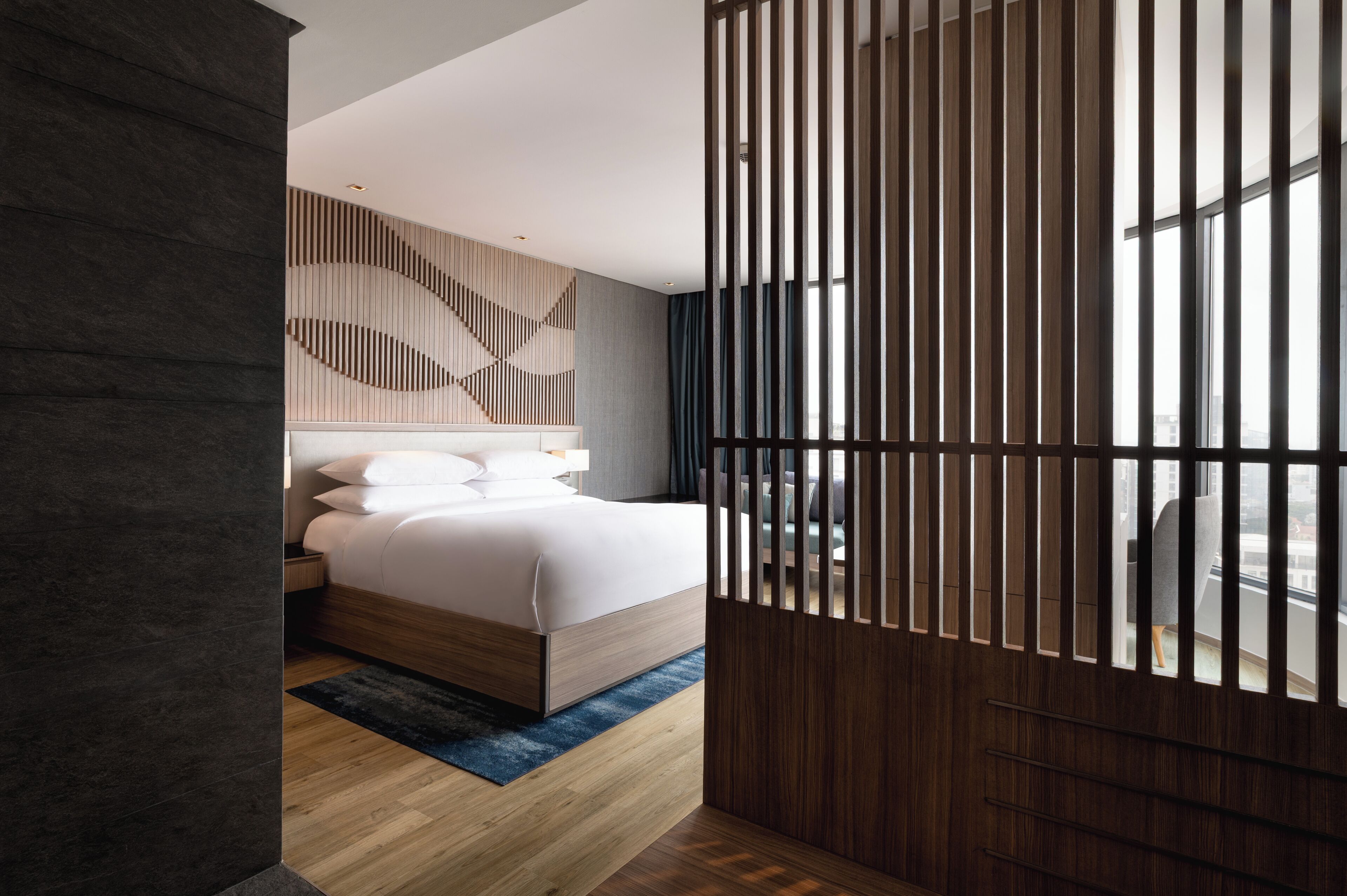 A modern hotel room with a bed, wooden panel wall, and a view through a window.