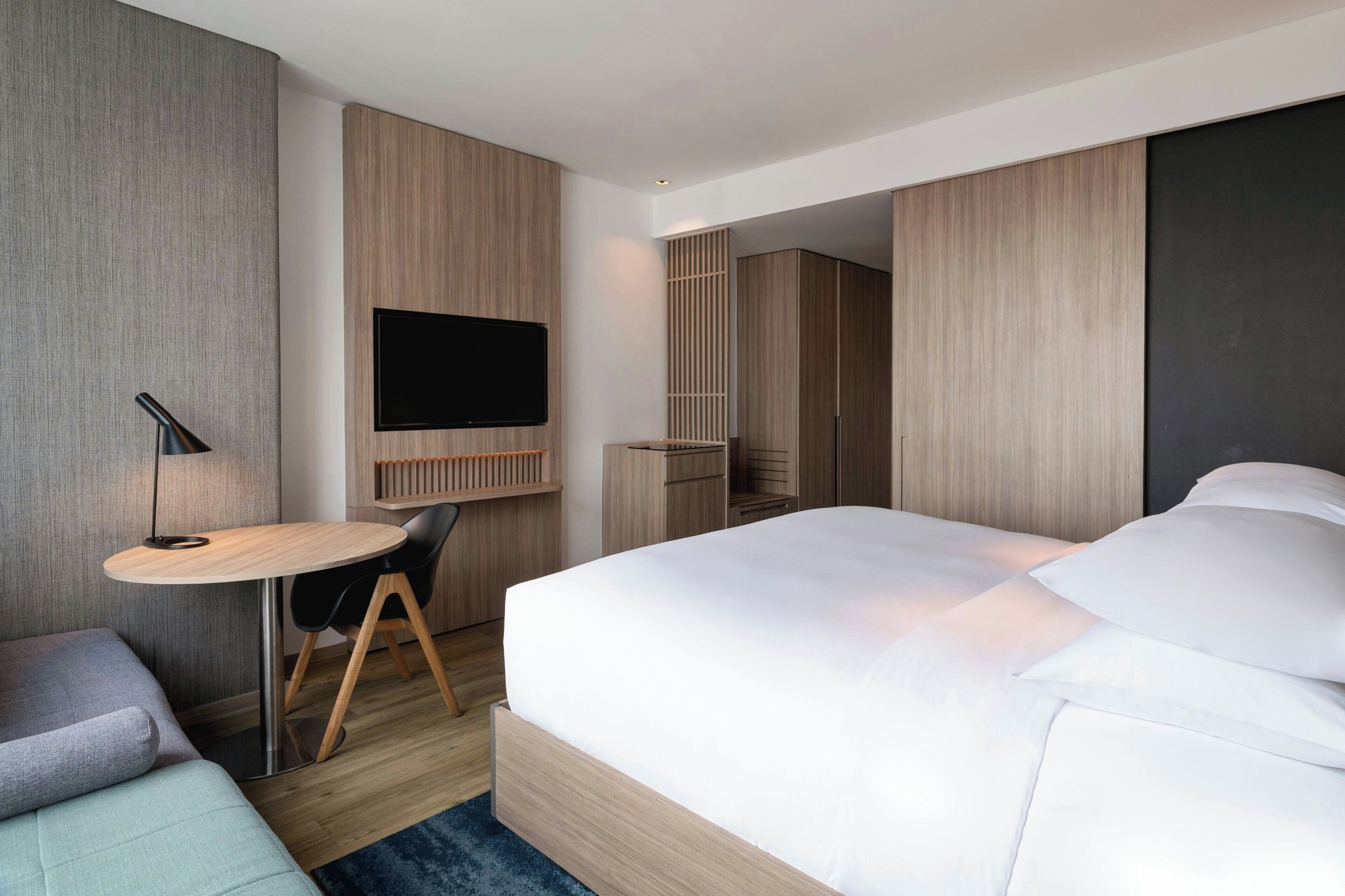 Una habitación de hotel moderna con una cama, un escritorio, una silla, un televisor y un armario.