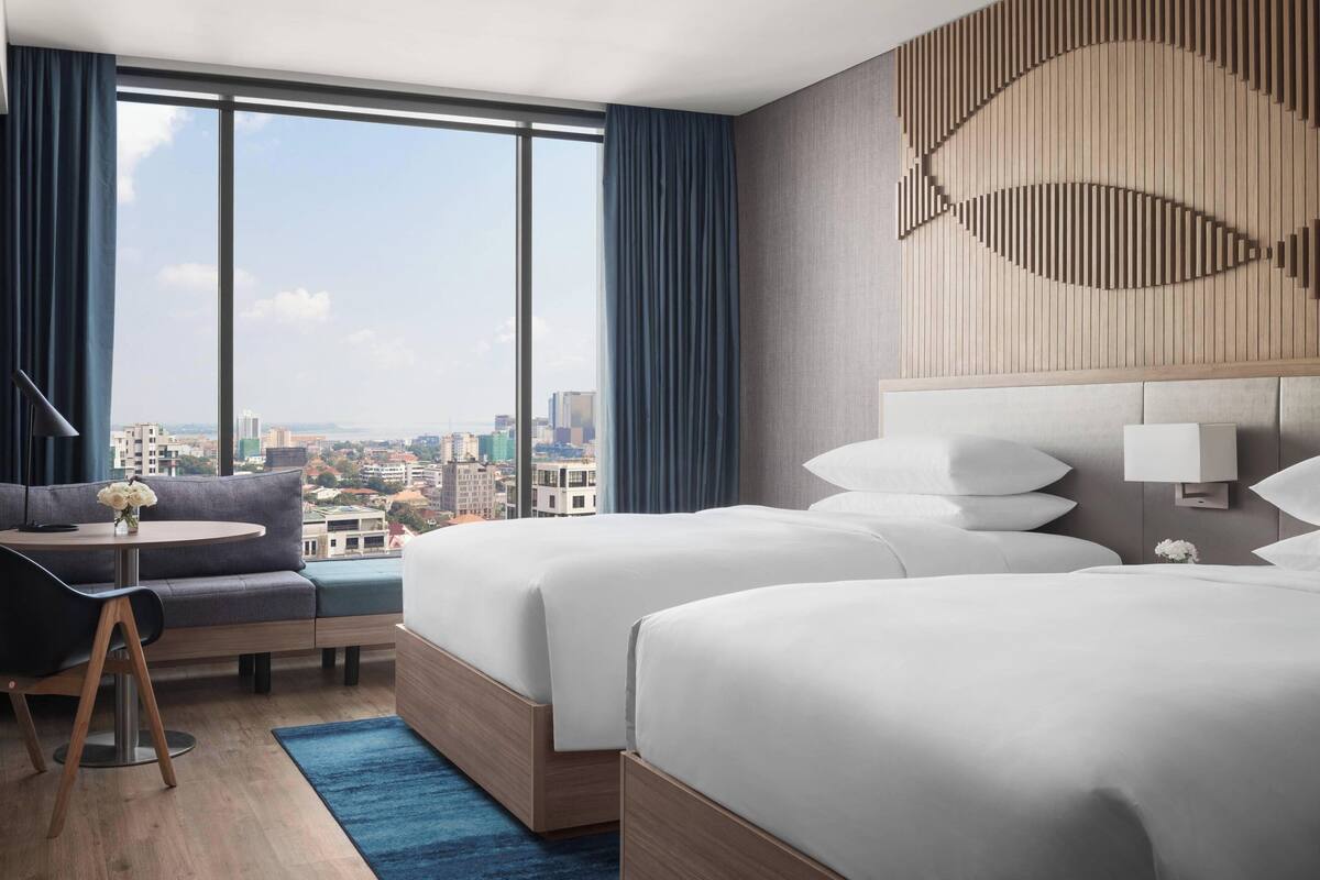 Habitación de hotel con dos camas, un sofá, una mesita con un florero y un ventanal con vistas a la ciudad.
