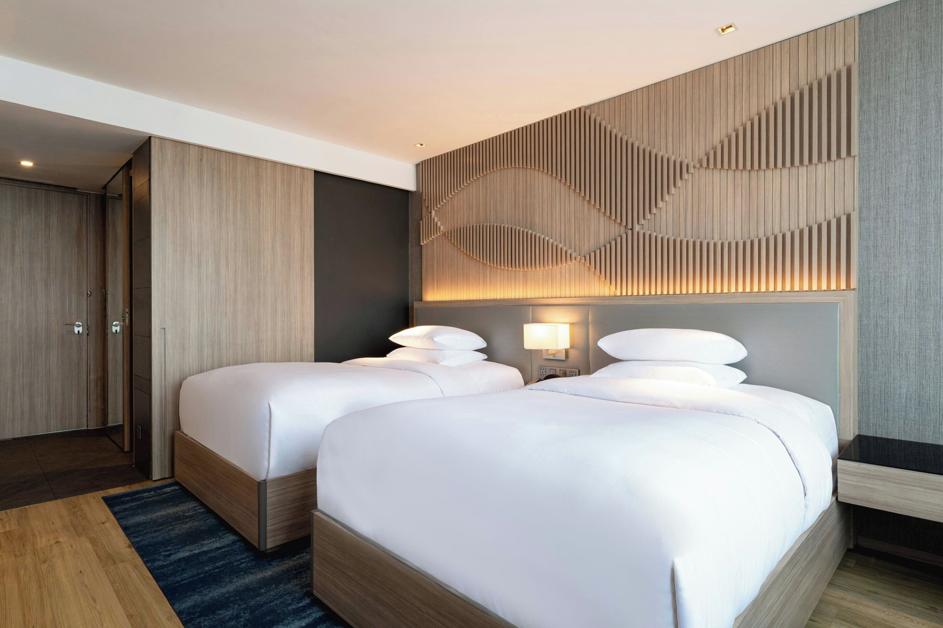 A hotel room with two beds, a wooden wardrobe, a wall-mounted light, and a patterned wall design.