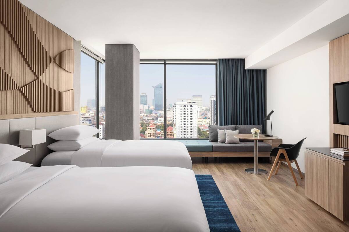 Una habitación de hotel moderna con dos camas, un escritorio, una silla, un televisor y un ventanal con vistas a la ciudad.