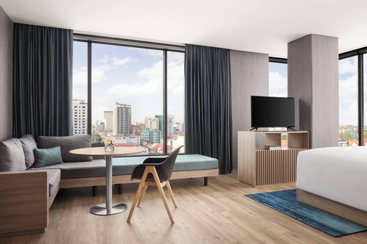 Una habitación de hotel moderna con un ventanal grande, un sofá, una mesa de comedor, un televisor y una cama.