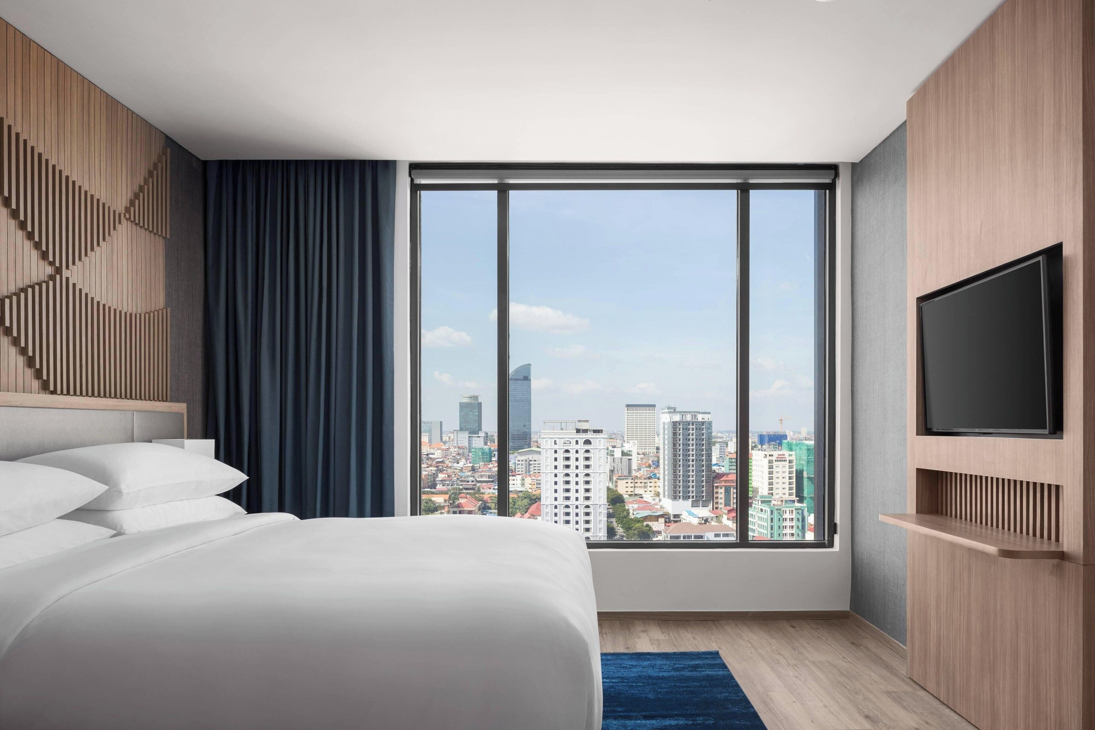 A modern hotel room with a large window, a bed, a TV, and a city view.