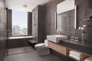 A modern bathroom with a bathtub, toilet, and sink. There is a large mirror above the sink and a window providing a view of buildings outside.