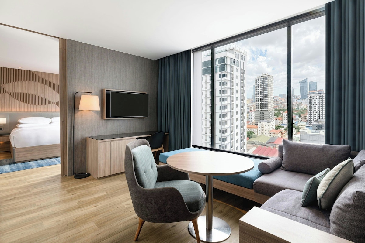Habitación de hotel moderna con cama, escritorio, televisor, sofá y vistas a la ciudad.