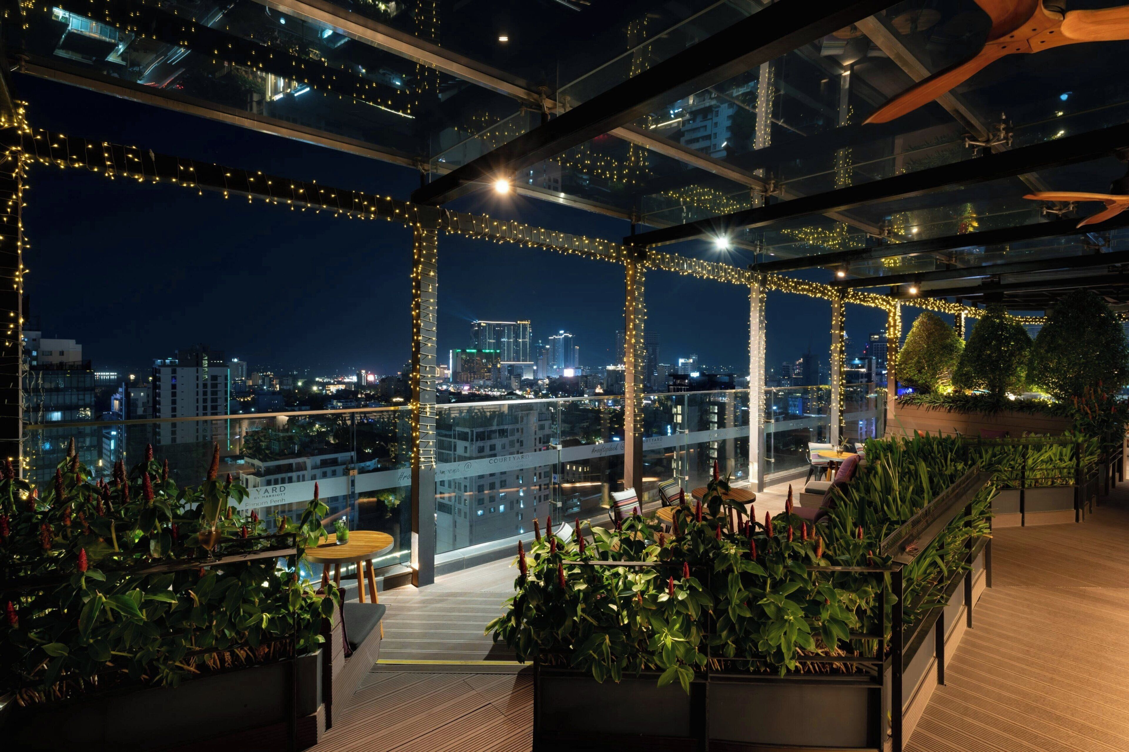 A rooftop terrace with glass railings, potted plants, and a city view at night.