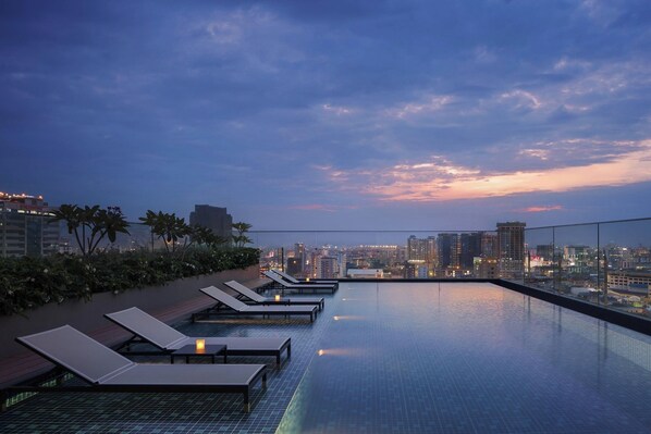 Piscina infinita en la azotea con sillones de descanso y vista panorámica del paisaje urbano al atardecer.
