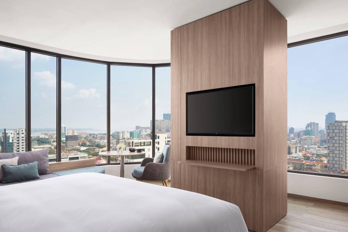 Una habitación de hotel moderna con una cama grande, un televisor montado y vistas a la ciudad a través de amplias ventanas.