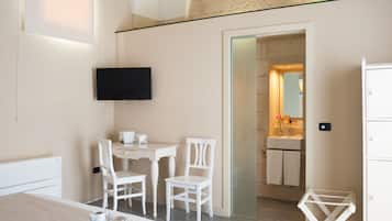 A hotel room with a bed, a desk, two chairs, a TV, a small table with a tray of pastries, and a bathroom visible through an archway.