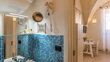 A bathroom with a blue-tiled wall, a hairdryer mounted on the wall, a sink with a mirror, and a table with chairs in the adjacent room.