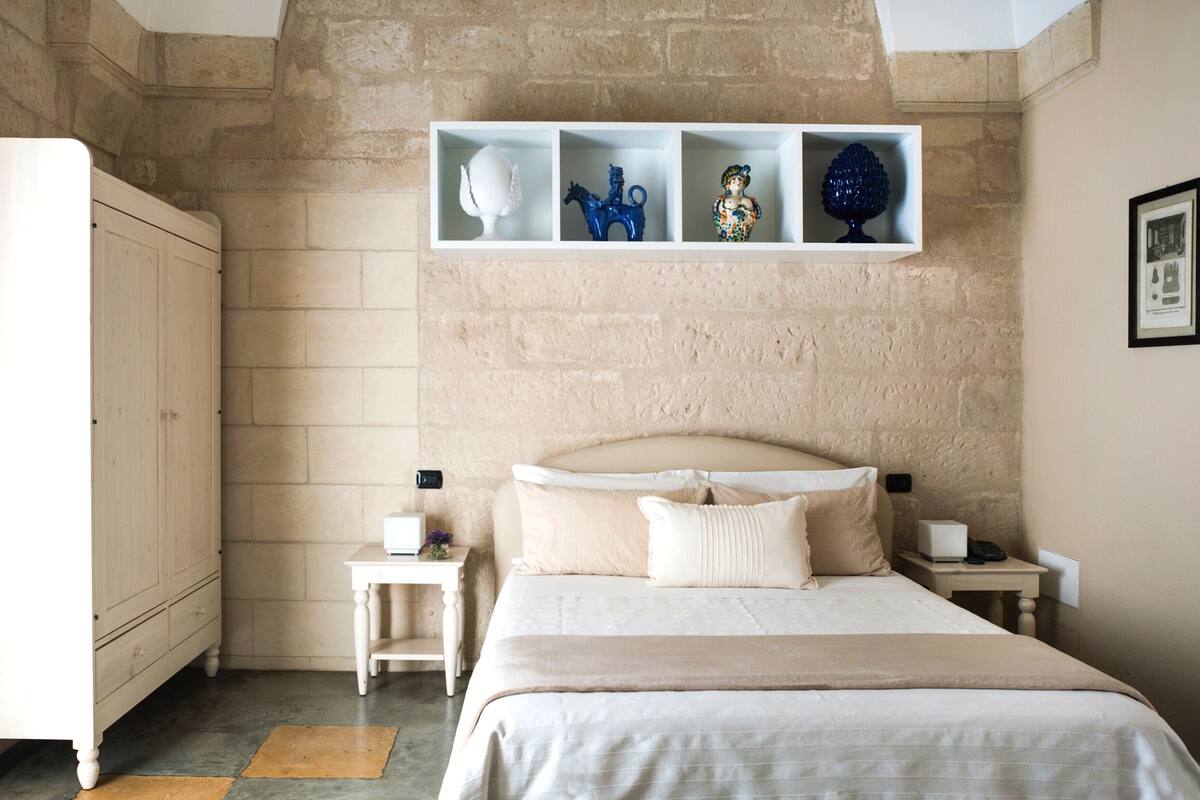A bedroom with a large bed, a wardrobe, a bedside table with a lamp, and a shelf with decorative items.