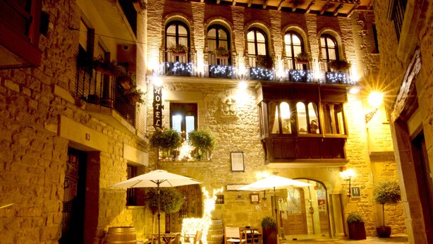 Edifici de pedra amb un rètol que diu “HOTEL” i un balcó amb plantes penjants. Hi ha taules i cadires a l'exterior sota ombrel·les.