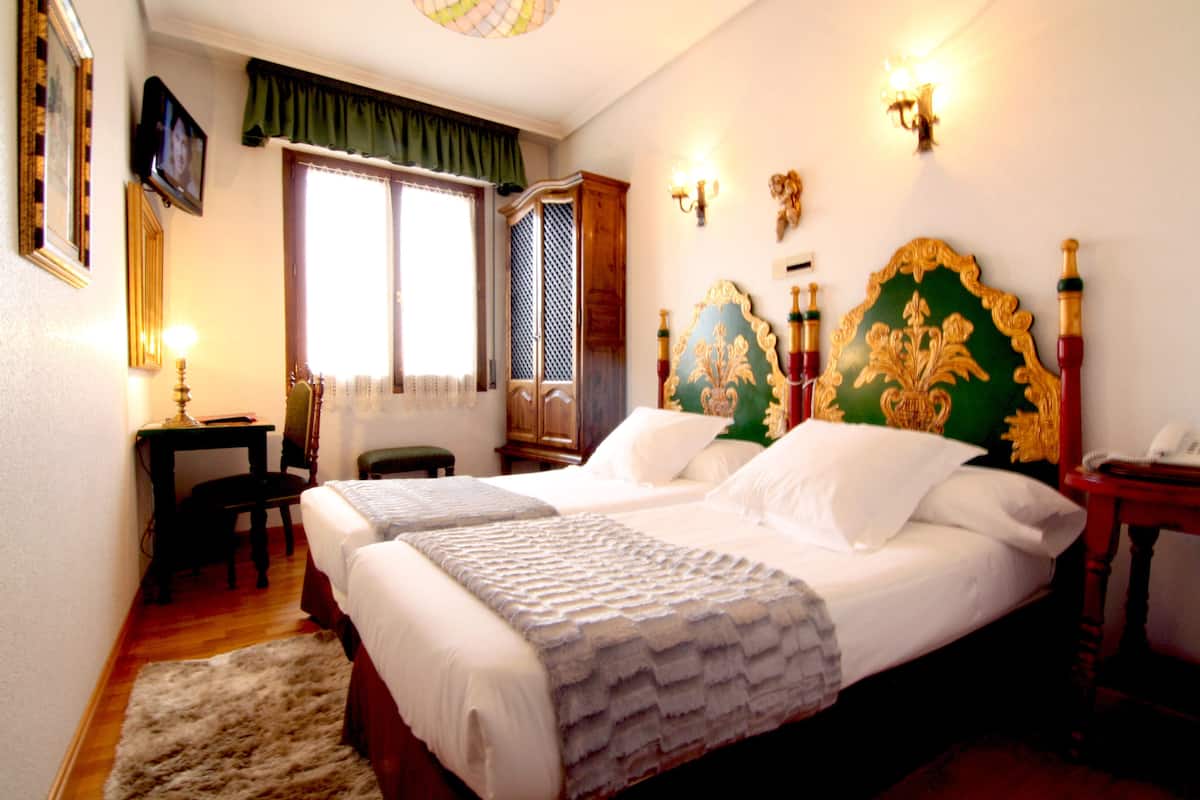 Una cama doble con un cabecero decorativo, un escritorio con silla, un televisor en la pared y una ventana con cortinas.