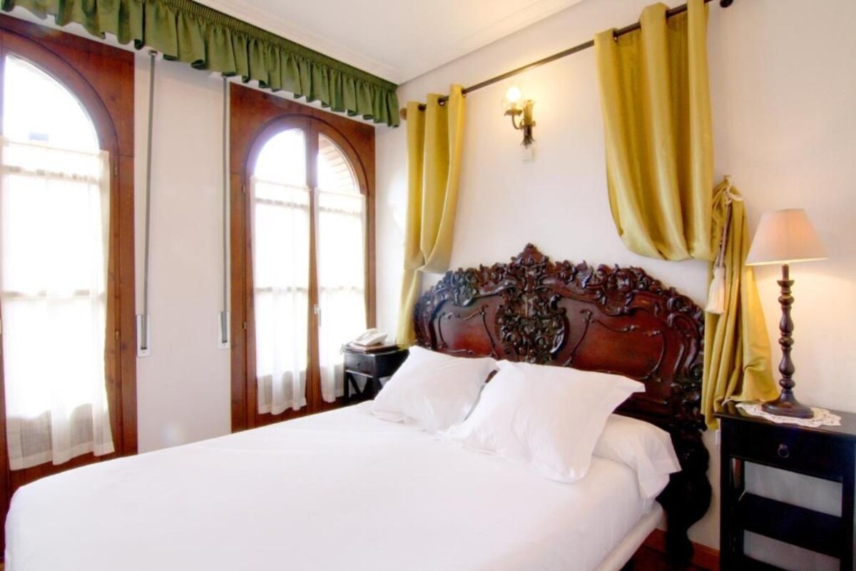 A hotel room with a large bed, a wooden headboard, two bedside tables, a lamp, and a window with curtains.