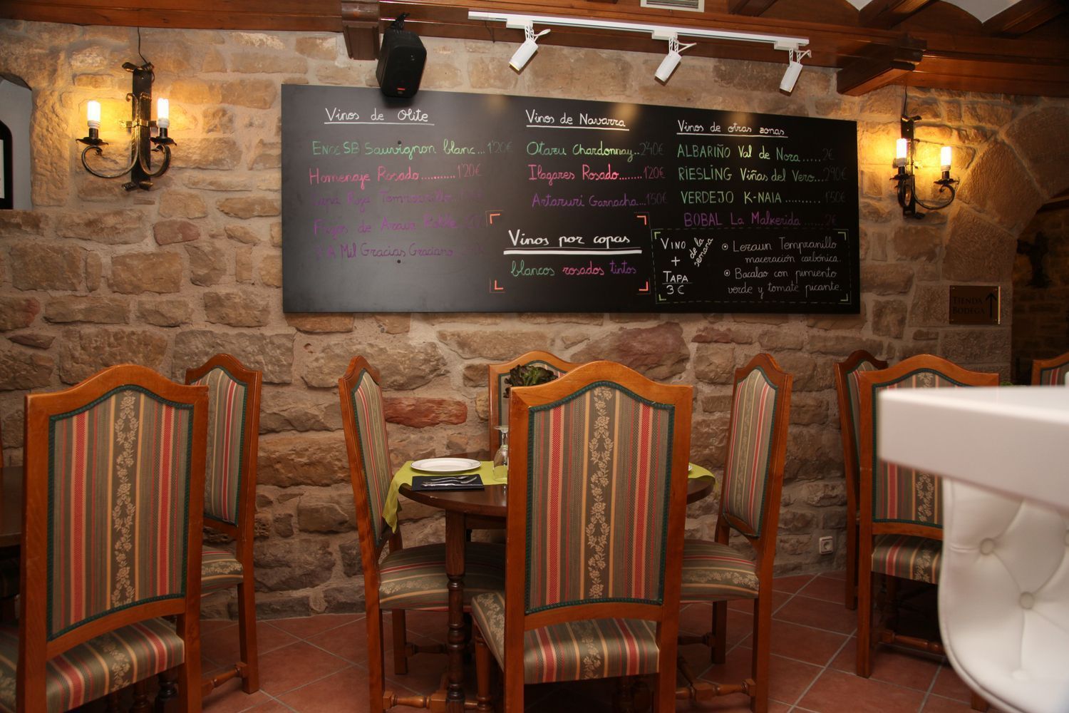 A restaurant with a stone wall, wooden chairs, and a chalkboard menu listing various wines and dishes.