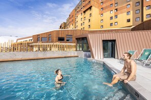 A swimming pool with two people, one sitting and one standing, surrounded by modern buildings. - Hôtel Club MMV Les Bergers (Huez)