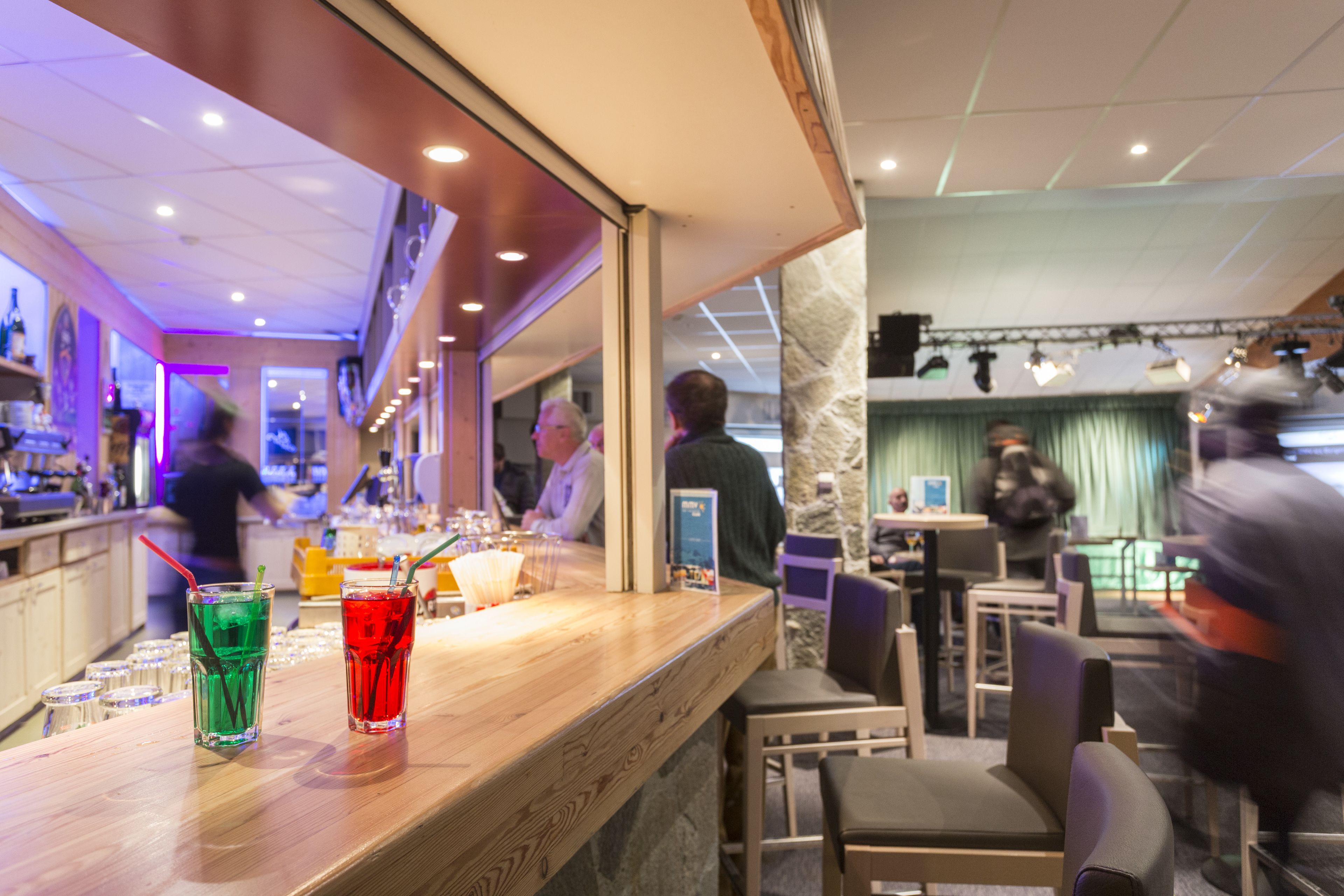 Un coin bar avec deux boissons posées sur le comptoir, un espace salon avec des tables et des chaises, et une personne à l’arrière-plan.
