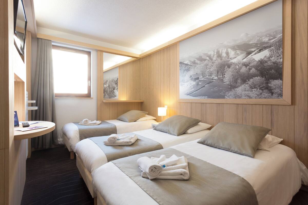 A hotel room with two beds, a bedside table, a window with curtains, a wall-mounted TV, and a large landscape painting.