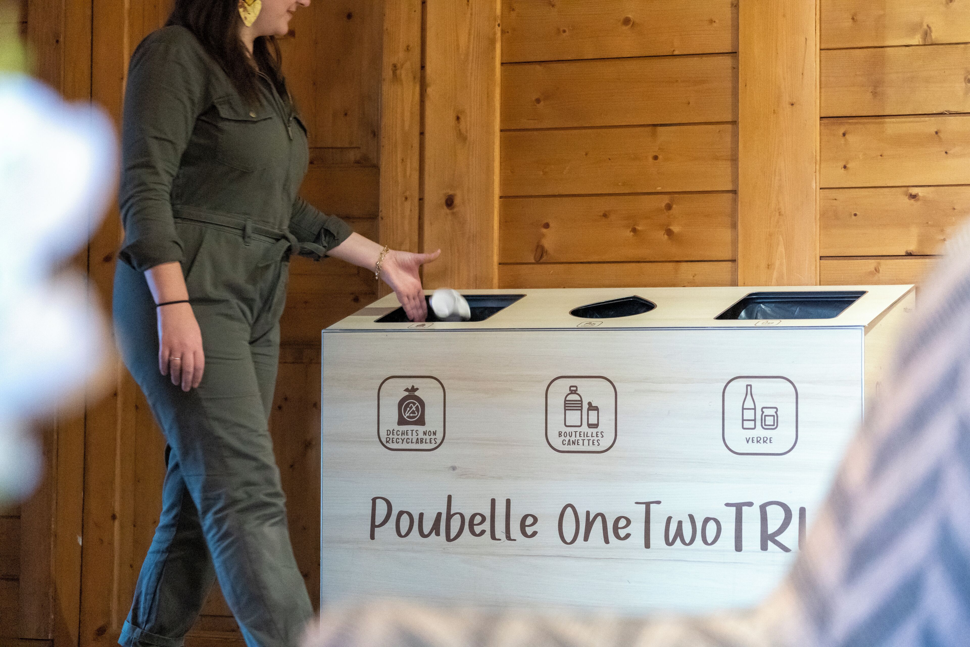 A woman placing a white object into a trash bin labeled "Poubelle OneTwoTRI" with recycling symbols for non-recyclable, glass, and bottle waste.