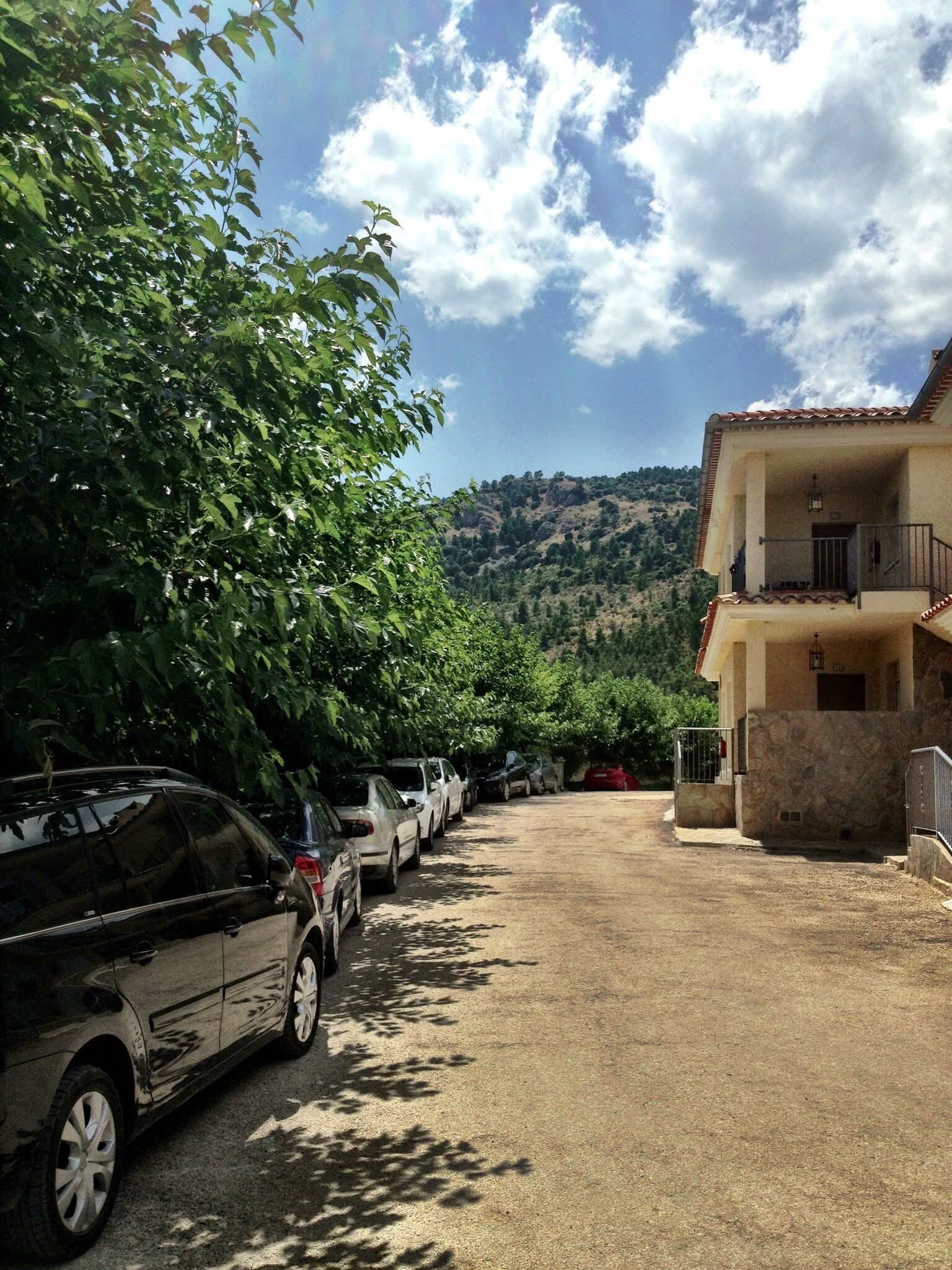 Un aparcamiento con coches, un edificio con balcón y una ladera al fondo.