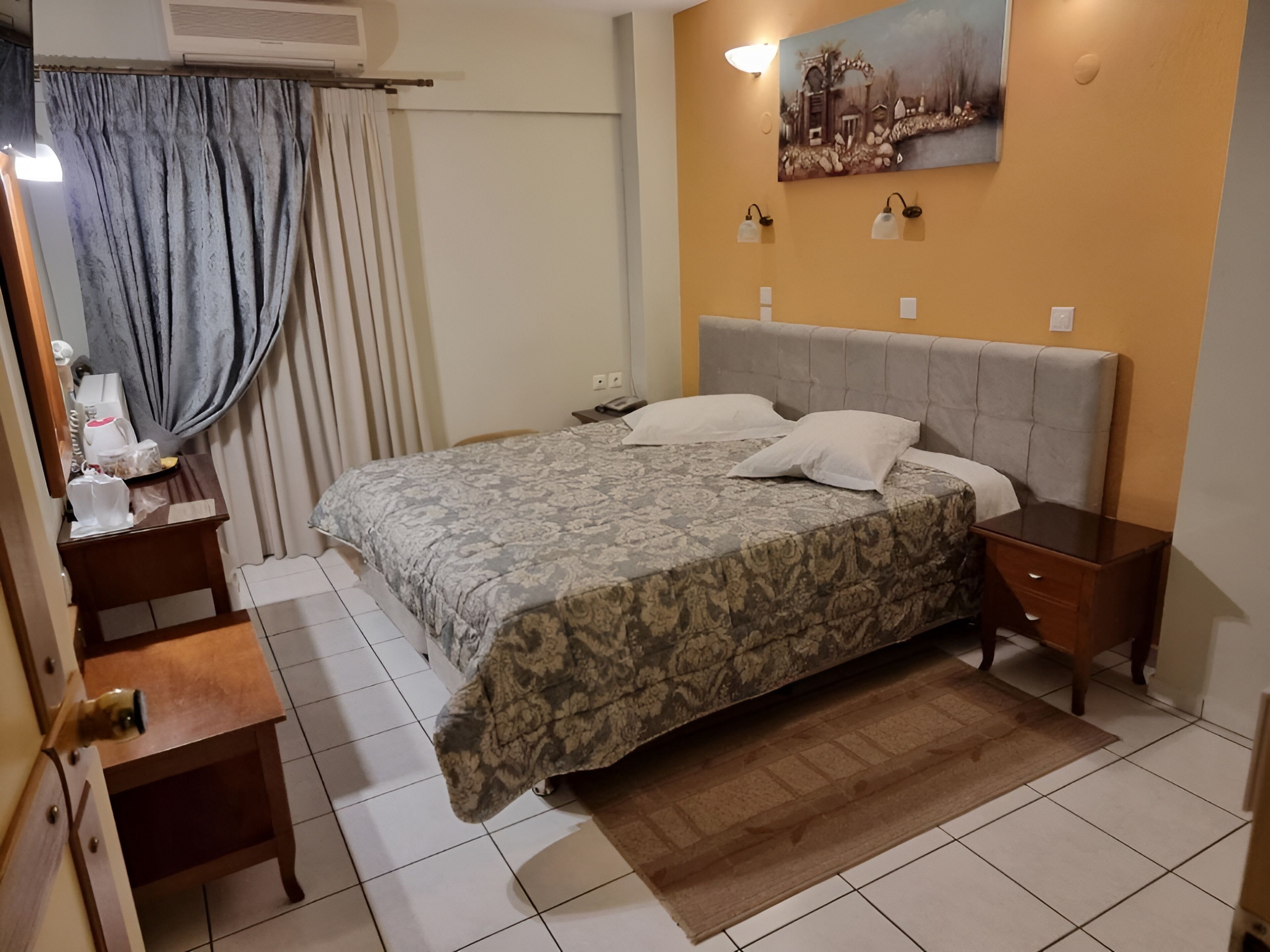 A hotel room with a bed, bedside tables, a desk, a chair, a window with curtains, and a painting on the wall.