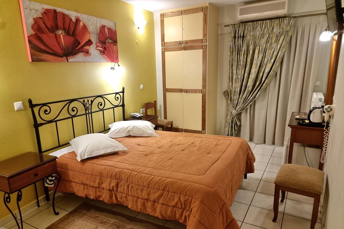 A bedroom with a bed, a nightstand, a chair, a desk with a telephone, and a painting on the wall.
