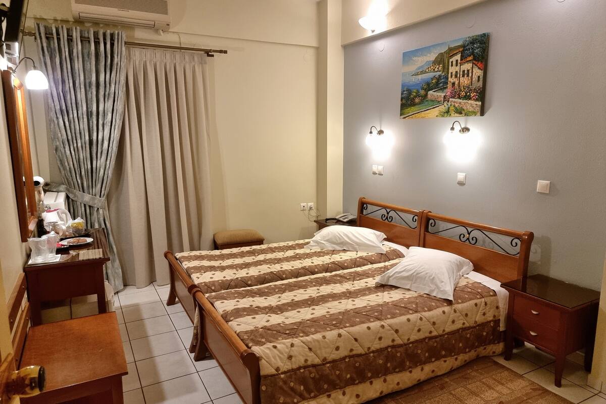 A hotel room with a bed, bedside tables, a desk, a chair, a curtain, and a painting on the wall.