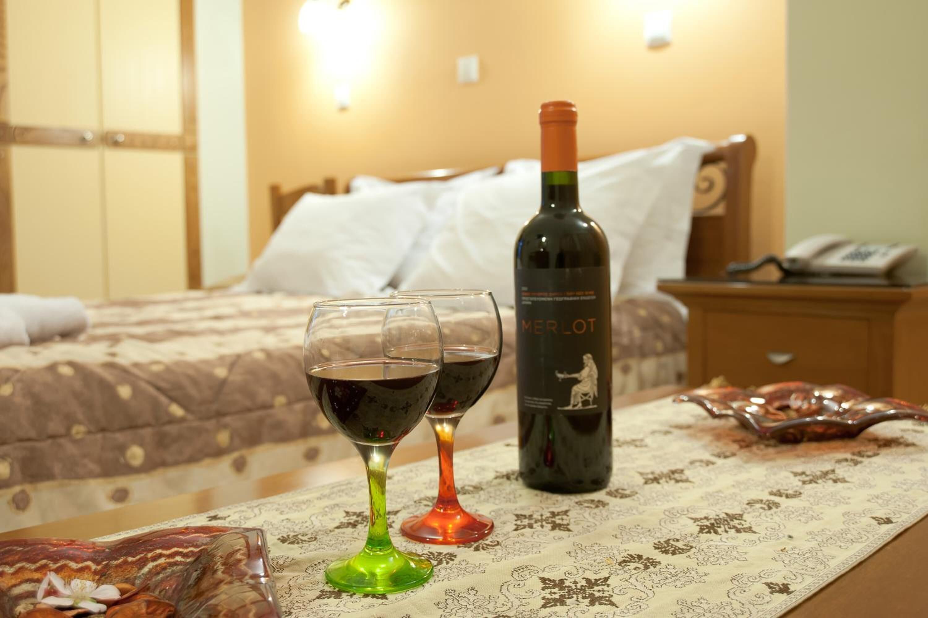 A hotel room with a bed, a bedside table with a bottle of Merlot wine and two glasses, and a patterned tablecloth.