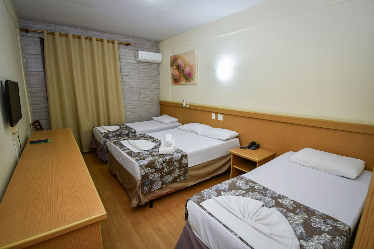 Quarto de hotel com duas camas de solteiro, uma escrivaninha de madeira, uma pequena mesa de cabeceira e ar condicionado fixado na parede.
