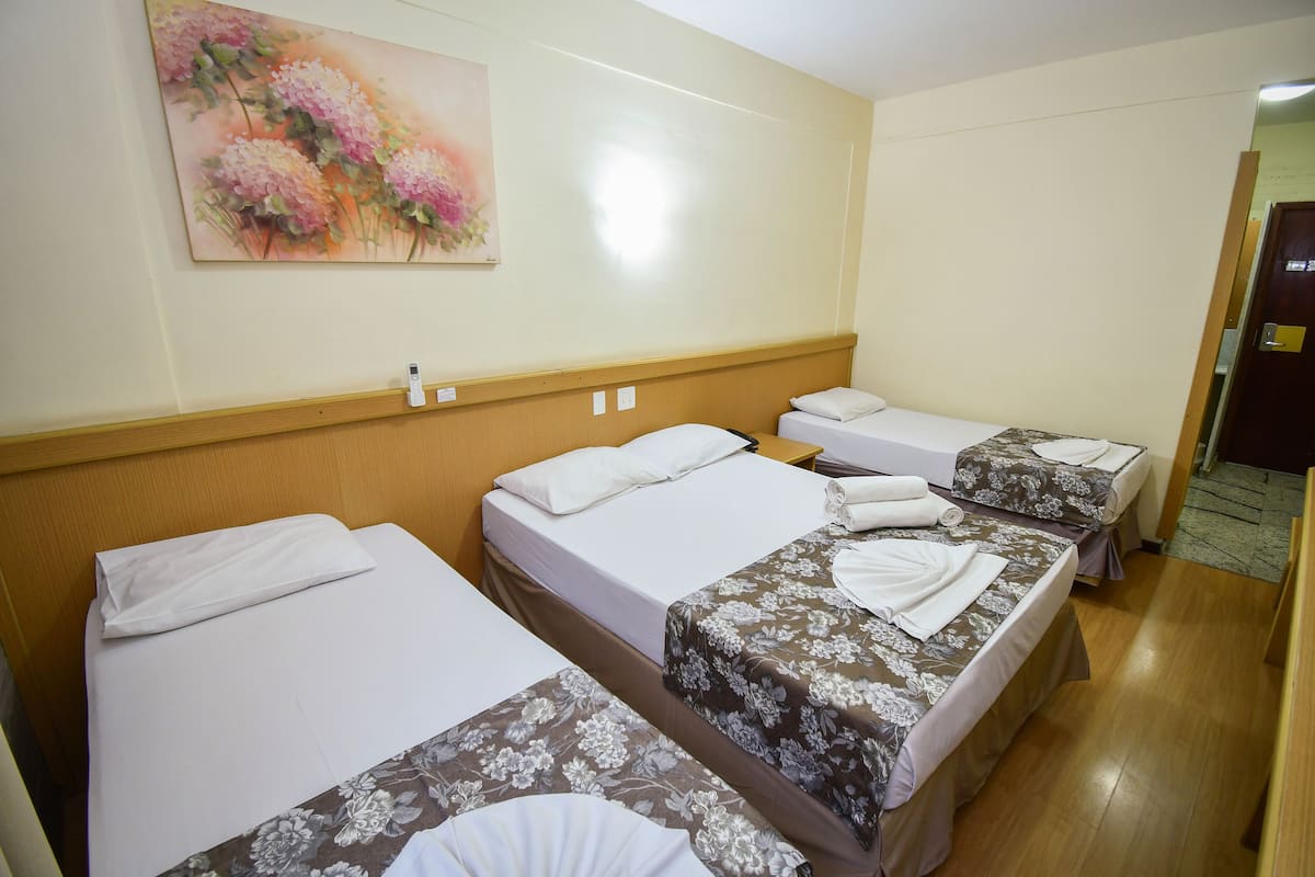 Quarto de hotel com três camas de solteiro, cada uma com colcha florida e almofadas brancas. Um quadro de flores está pendurado na parede acima das camas.