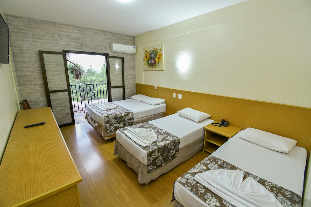 Quarto de hotel com três camas de solteiro, uma escrivaninha de madeira, varanda com vista e um quadro na parede.