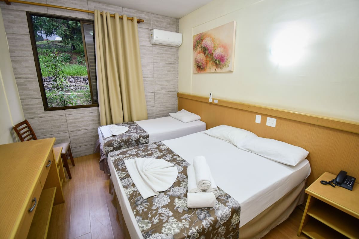 Quarto de hotel com duas camas, uma escrivaninha de madeira, uma cadeira, janela com vista para vegetação e um quadro na parede.