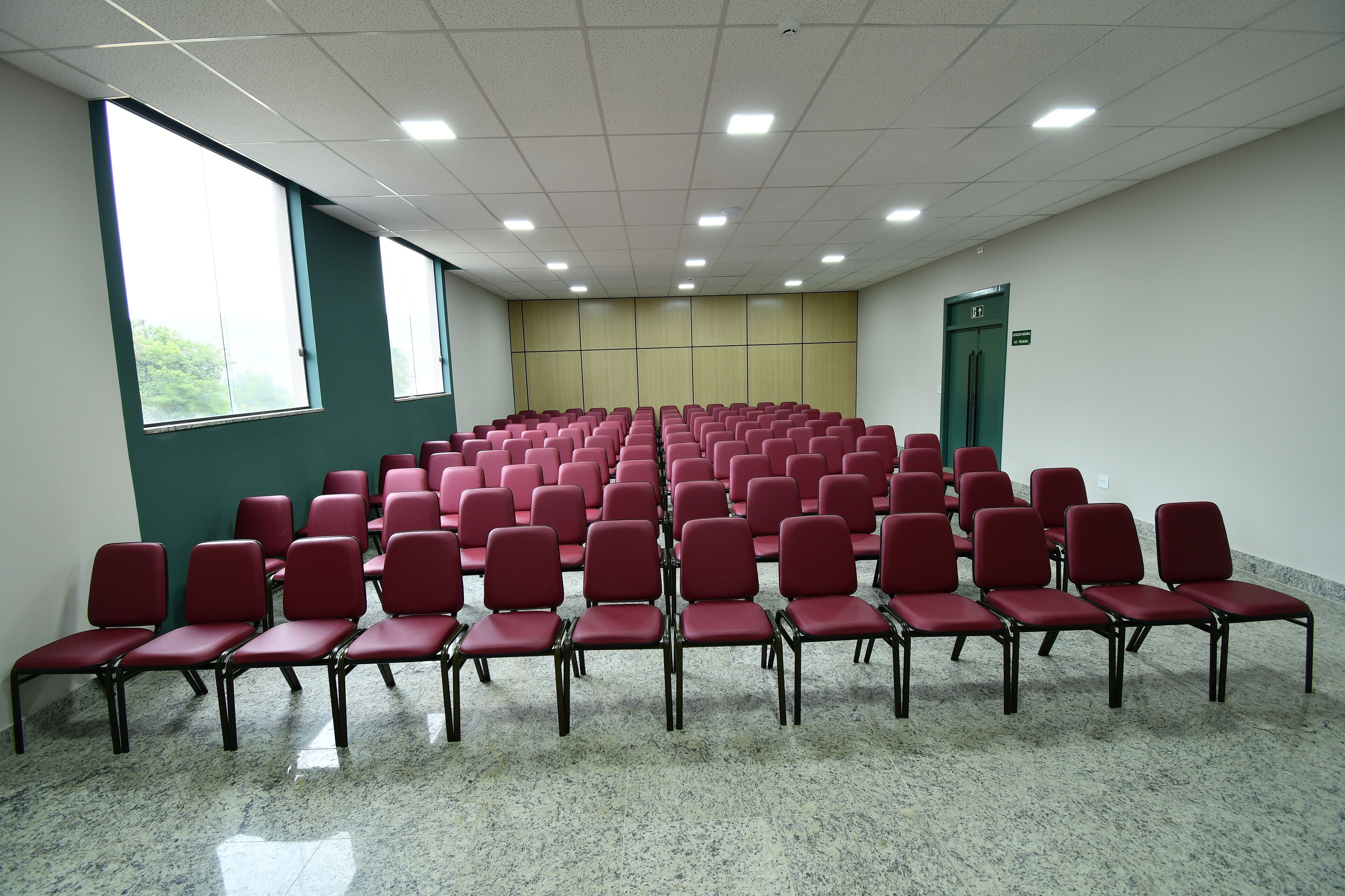 A conference room with rows of red chairs, large windows, and a green door.