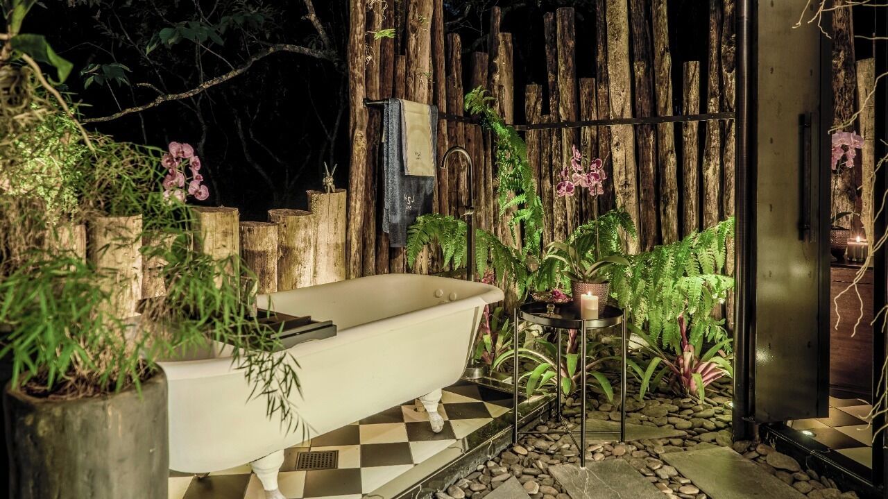 Una bañera en un entorno rústico al aire libre, con troncos de madera, plantas y un suelo de cuadros.