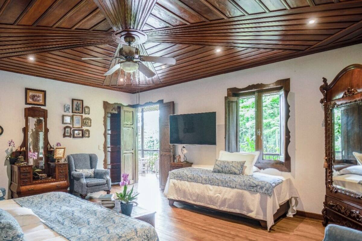Une chambre avec deux lits, un ventilateur de plafond, des poutres apparentes, un grand miroir et une vue sur la verdure environnante.