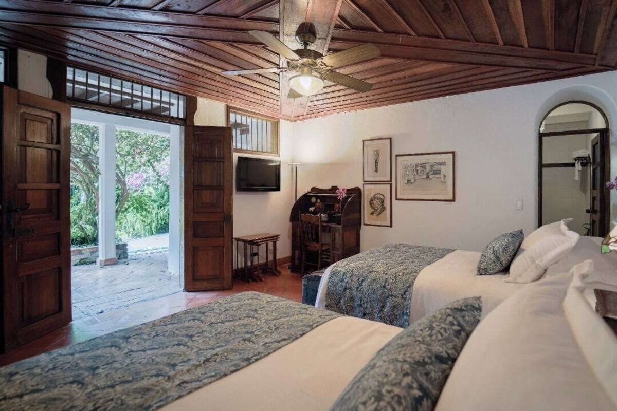 Une chambre avec deux lits, un ventilateur de plafond, une porte en bois, un bureau et une lampe.